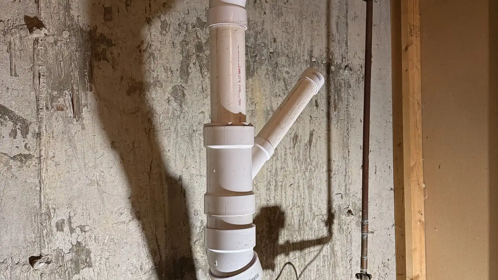 A white PVC drain pipe with a wye fitting and capped branch installed against an unfinished concrete wall in a basement.